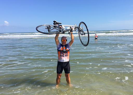Angel Kowalski standing in the ocean shore raising his bicycle in the air after a ride