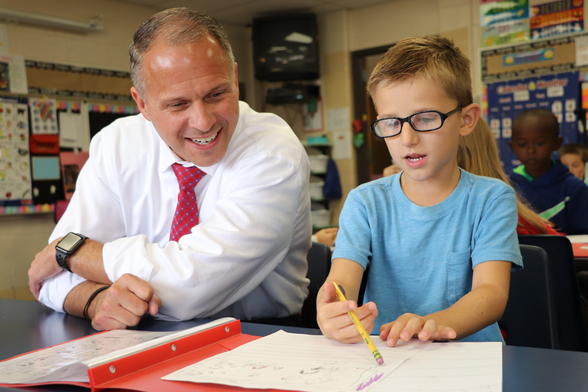 Boy sitting at classroom table shows superintendent his drawing