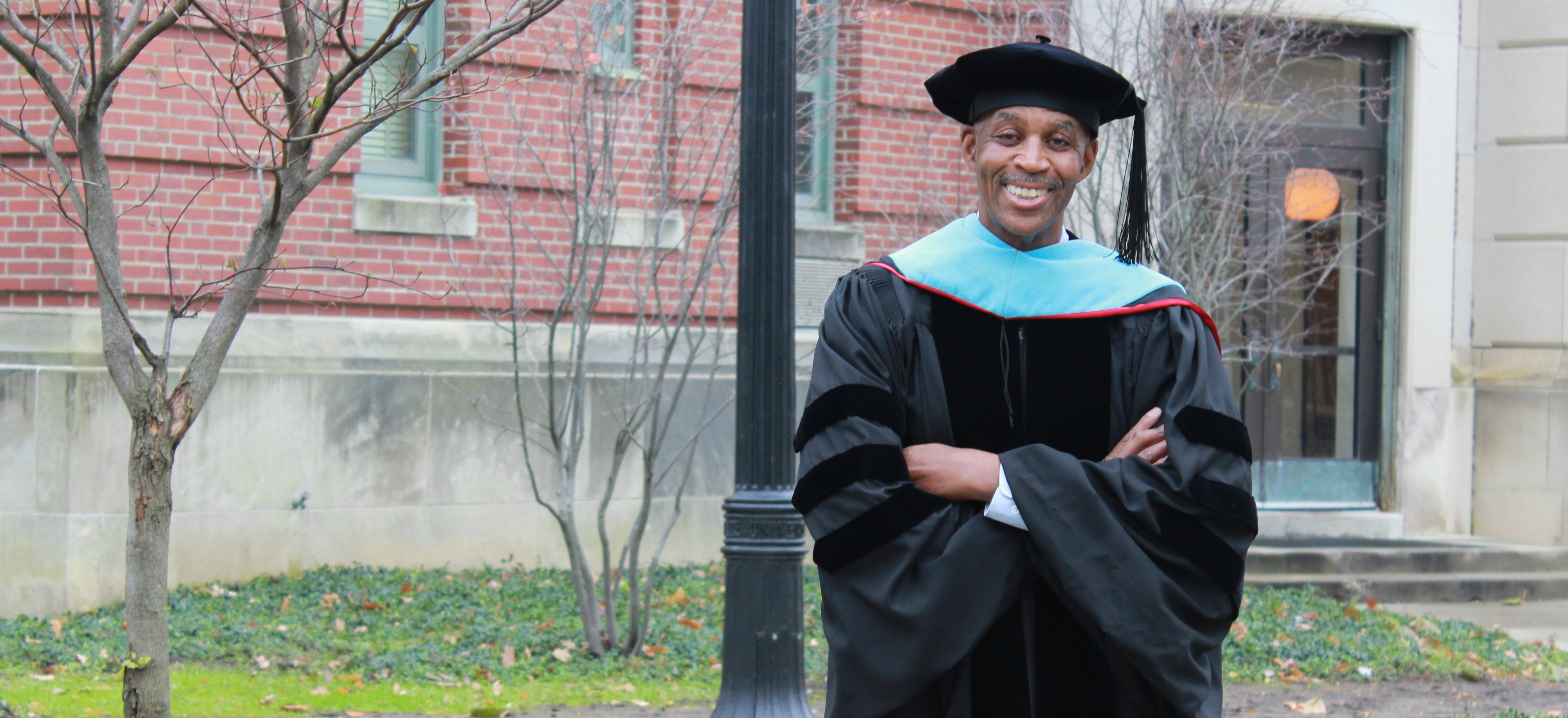 Keith Bell poses in his graduation cap and gown in front of Ramseyer Hall, where he conducted research for his Doctor of Education degree.