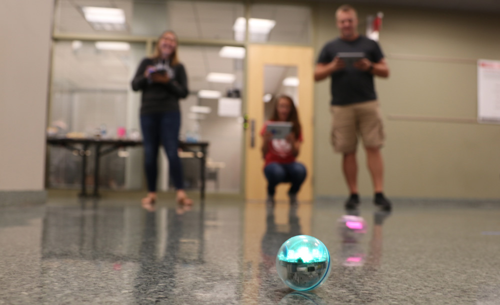 Three people in the background make an app-controlled ball move along the floor