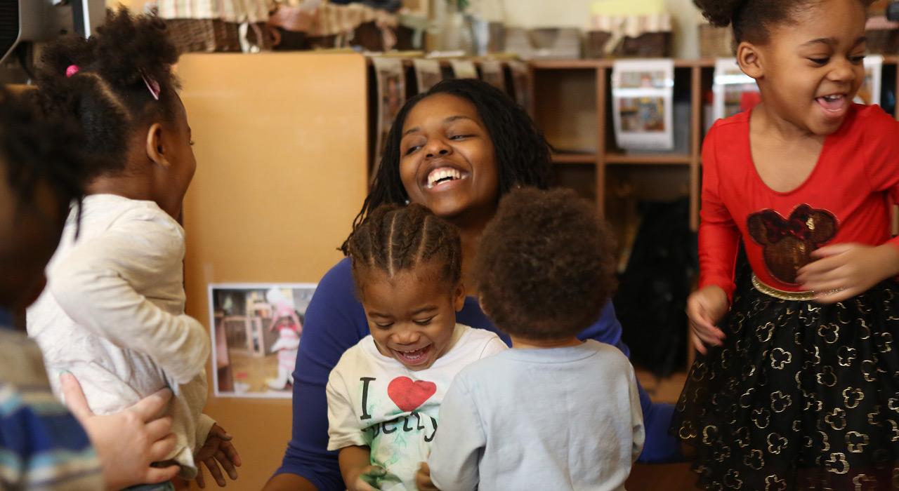 preschool teacher laughing as young children play around her