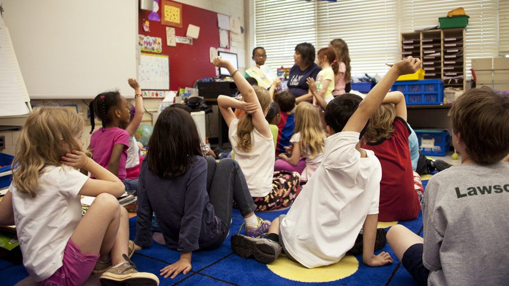 A classroom of elementary students sit on the floor listening as a teacher reads a book