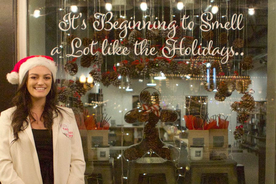 woman in Santa hat stands in front of a candle store window display she designed