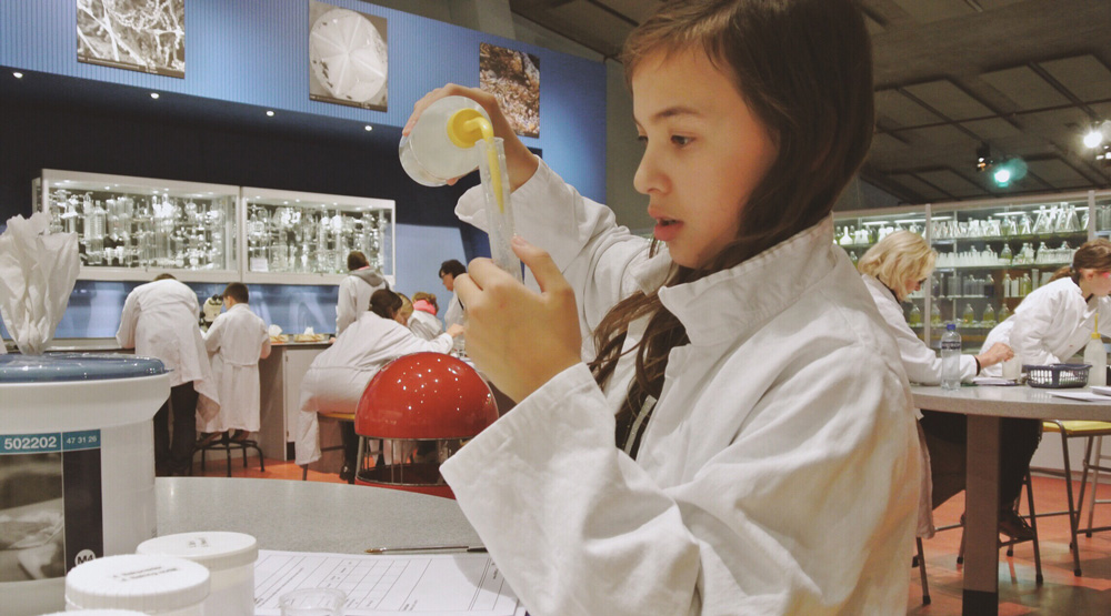 Girl wearing white lab coat pours substance into a test tube