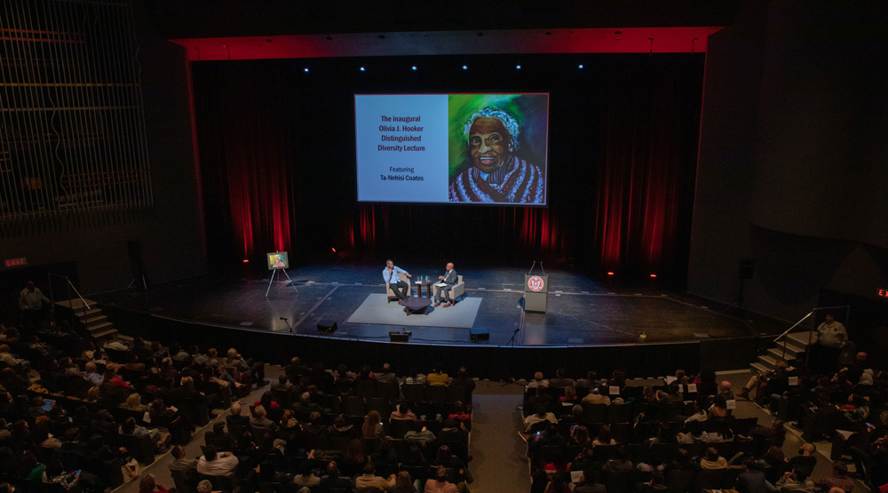 Two men on stage talking with hundreds listening in from auditorium seats