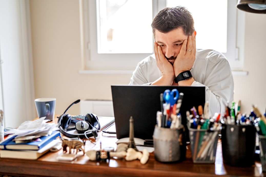 frustrated man rests hands on chin working on laptop at messy home office