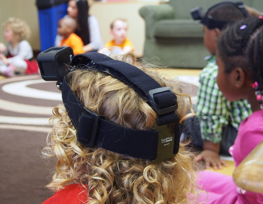 A preschool-aged girl with curly hair wears a video camera to record her classroom perspectives