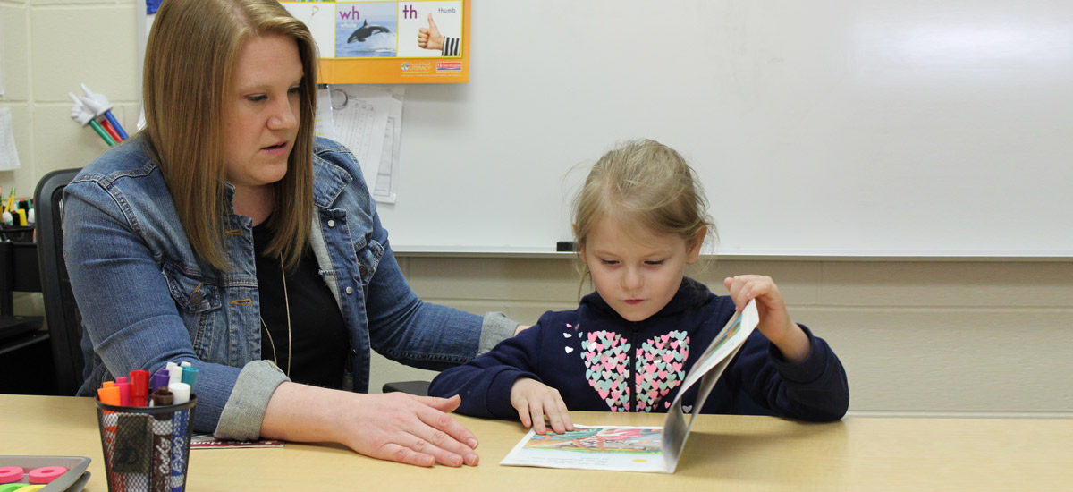 Girl reads book to woman in classroom