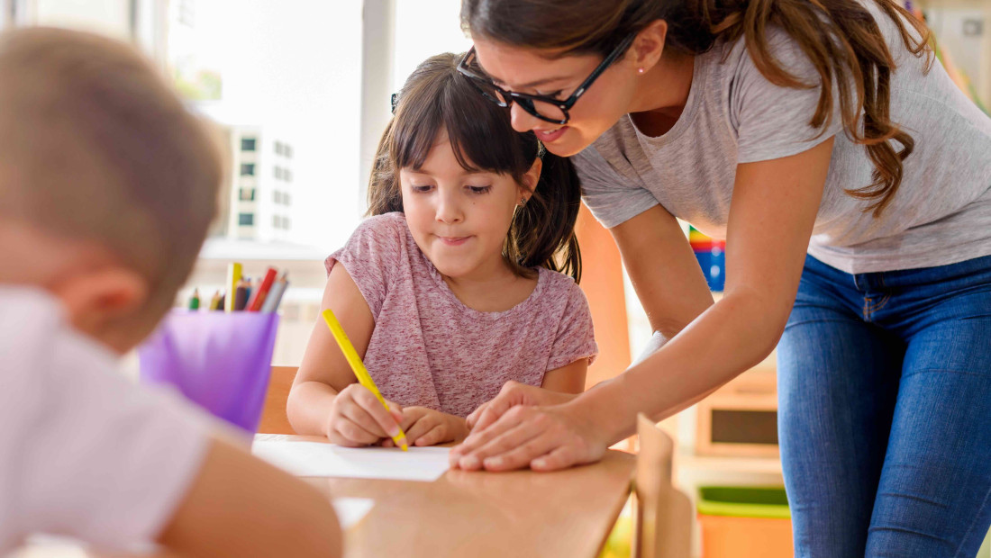 Teacher assisting a young student with a writing assignment in a classroom 