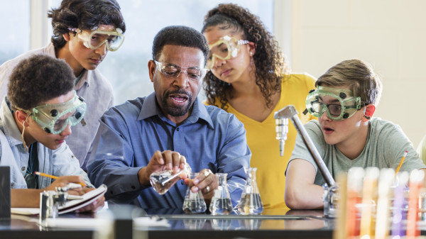 Teacher sitting at a chemistry table with students wearing safety goggles conducting an experiment