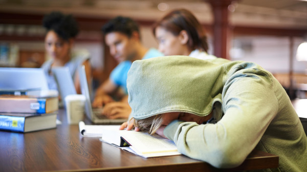 college student in a grenn hoodie studying in a library with their head down on the table and hood up