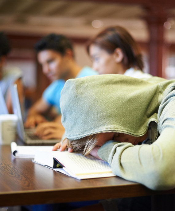 college student in a grenn hoodie studying in a library with their head down on the table and hood up