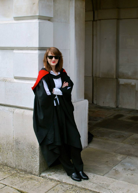 Woman wearing doctoral regalia and sunglasses leans against the archway on a building