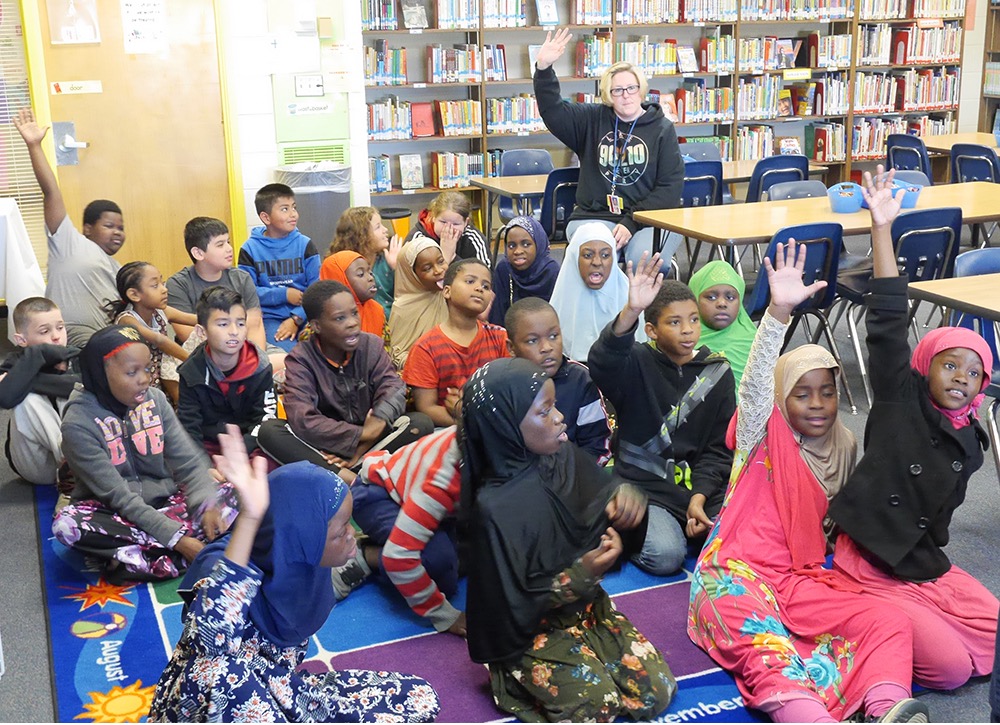 classroom of students sitting on floor raising their hands